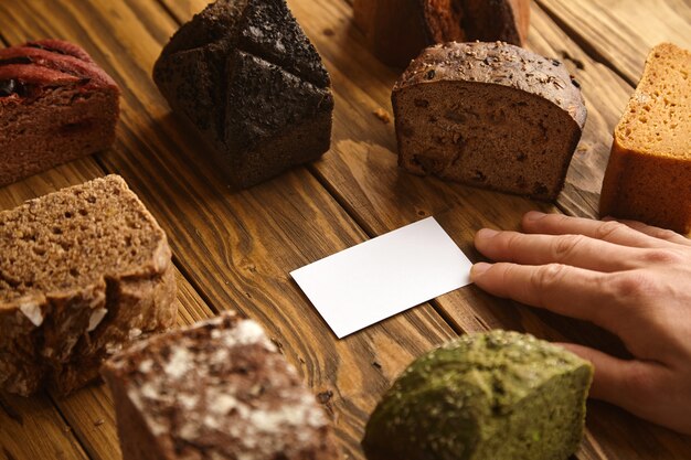 Hand nimmt leere Visitenkarte des professionellen handwerklichen Bäckers präsentiert in der Mitte vieler gemischter alternativer gebackener exotischer Brotproben über rustikalem Holztisch