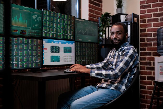 Handelsagent einer afroamerikanischen Investmentgesellschaft sitzt am Schreibtisch mit mehreren Displays und blickt in die Kamera. Finanzmakler im Büro einer Handelsagentur mit Echtzeitgrafiken auf Monitoren.