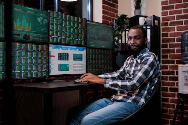 Handelsagent einer afroamerikanischen Investmentgesellschaft sitzt am Schreibtisch mit mehreren Displays und blickt in die Kamera. Finanzmakler im Büro einer Handelsagentur mit Echtzeitgrafiken auf Monitoren.