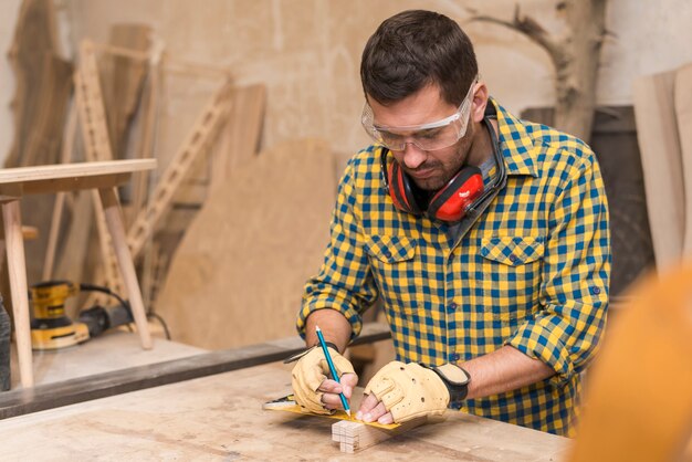 Handwerkerhände in den Schutzhandschuhen, die Holzklotz mit Machthaber und Bleistift messen