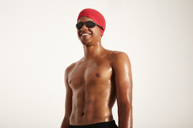 Happy Fit muskulös lächelnder afroamerikanischer Schwimmer mit nasser Haut, die rote Kappe und schwarze Schutzbrille trägt, die weg schauen