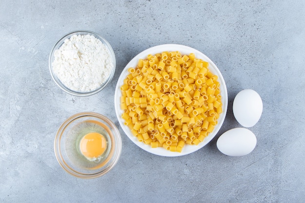 Haufen roher Pipetten-Rigate-Nudeln in einer weißen Schüssel mit Eiern und Mehl.