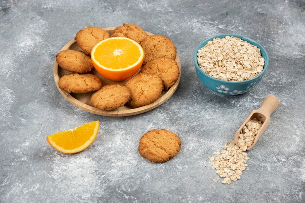 Haufen von Keksen mit Orange auf Holzbrett und Haferflocken in einer Schüssel.