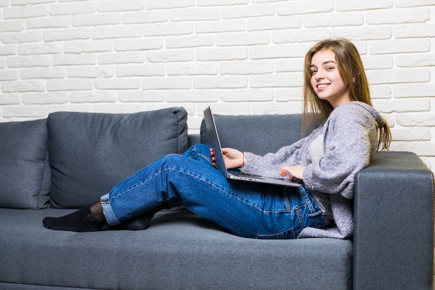 Haus-, Technologie- und Internetkonzept - beschäftigtes junges Mädchen, das auf der Couch mit Laptop-Computer zu Hause liegt