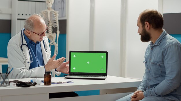 Hausarzt zeigt auf Laptop-Display mit Greenscreen, Treffen bei Kontrollbesuch. Blick auf den leeren Copyspace-Hintergrund mit isolierter Mockup- und Chroma-Key-Vorlage. Stativaufnahme.