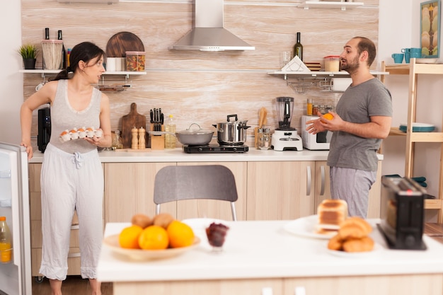 Hausfrau holt eier aus dem kühlschrank und trägt pyjamas, um das frühstück zu kochen. ehemann unterhält sich mit seiner frau, während sie das morgenessen kocht