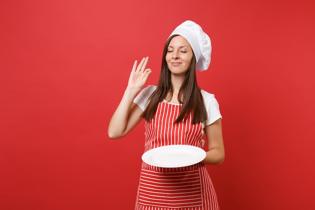 Hausfrau köchin oder bäckerin in gestreifter schürze, weißes t-shirt, haubenköche isoliert auf rotem wandhintergrund. frau hält leere leere runde platte mit platz für essen. mock-up-kopierraumkonzept