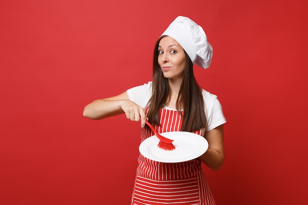 Hausfrau köchin oder bäckerin in gestreifter schürze, weißes t-shirt, haubenköche isoliert auf rotem wandhintergrund. frau hält weiße platte mit bürste zum abwaschen. mock-up-kopierraumkonzept.