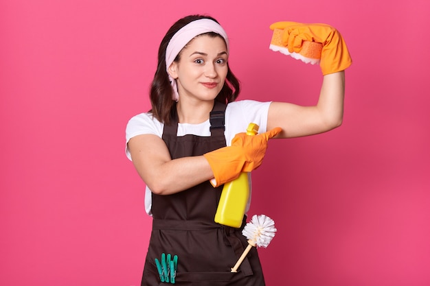 Haushälterin trägt weißes T-Shirt und braune Schürze und hält Schwamm in der Hand