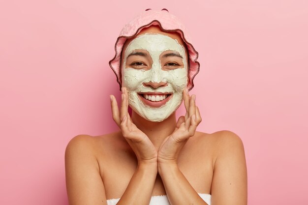 Hautpflege für alle Altersgruppen. Glückliche asiatische Dame mit schälender Tonmaske im Gesicht, hat Schönheitsbehandlungen, sieht angenehm aus, berührt Wangen, trägt Duschhaube