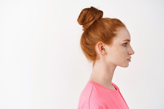 Hautpflege- und Make-up-Konzept Profil einer jungen Frau mit natürlichem rotem Haar, das in einem unordentlichen Brötchen gekämmt ist und nach rechts steht, auf weißem Hintergrund Gesichtsbehandlung