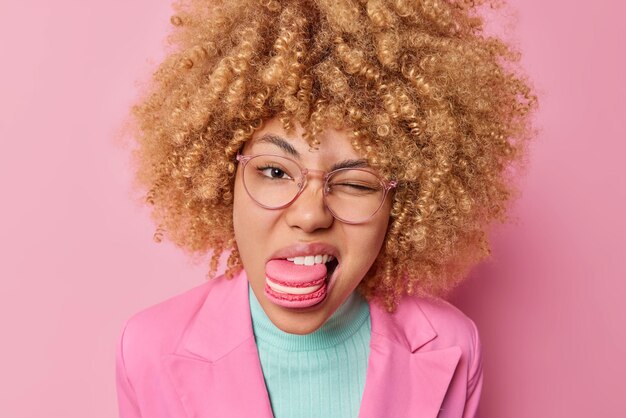 Headshot der hübschen Frau mit buschigen Locken hält köstlichen leckeren Donut im Mund zwinkert Auge trägt eine transparente Brille und eine formale Jacke, die über rosa Hintergrund isoliert ist Zuckerdiät oder Sucht