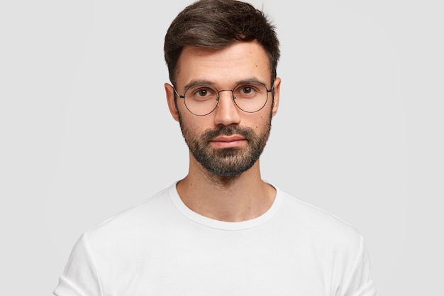 Headshot eines gutaussehenden männlichen Freiberuflers mit ansprechendem Aussehen, hat dunklen Bart und Schnurrbart, sieht direkt mit ernstem Blick aus, trägt weiße Freizeitkleidung. Einfarbig. Gesichtsausdrücke.