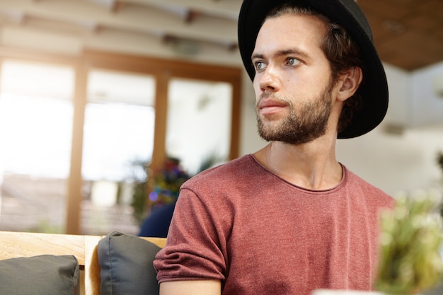 Headshot eines hübschen jungen Models mit flockigem Bart in stilvoller Kopfbedeckung und T-Shirt mit hochgekrempelten Ärmeln, die wegschauen, während sie drinnen posieren