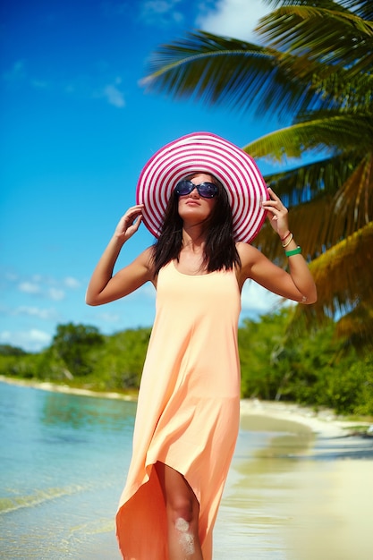 Heiße Schönheit im bunten sunhat und im Kleid gehend nahe Strandozean am heißen Sommertag nahe Palme
