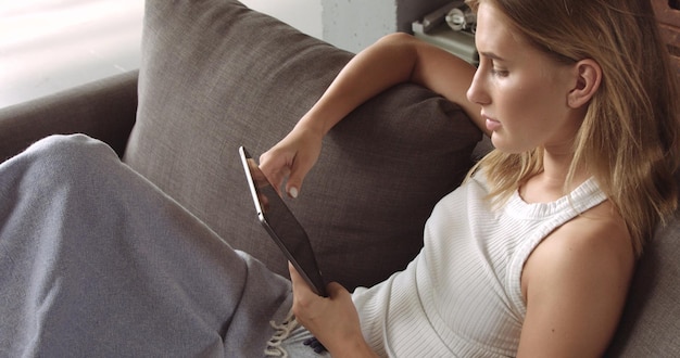 Heißes blondes Model liest auf ihrem Tablet, während es auf einer dunkelgrauen Couch sitzt, die mit einem weichen Überwurf bedeckt ist