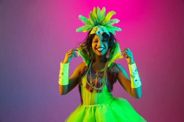 Hell. Schöne junge Frau im Karneval, stilvolles Maskeradenkostüm mit Federn, die auf Gradientenwand in Neon tanzen. Konzept der Feiertagsfeier, der festlichen Zeit, des Tanzes, der Party, des Spaßes.