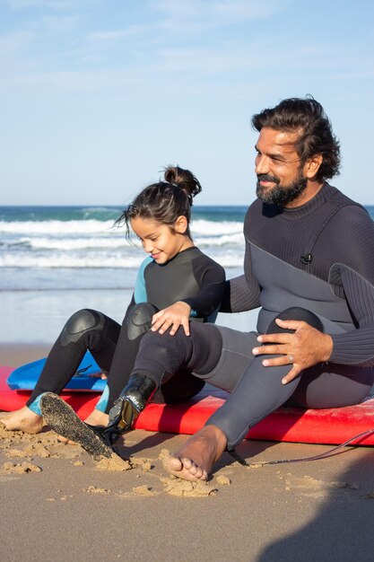 Heller Vater mit mechanischem Bein mit Tochter am Strand. Mitte erwachsener Mann und kleines dunkelhaariges Mädchen sitzen auf Surfbrettern und ruhen sich nach dem Training aus. Familie, Freizeit, aktives Lifestyle-Konzept