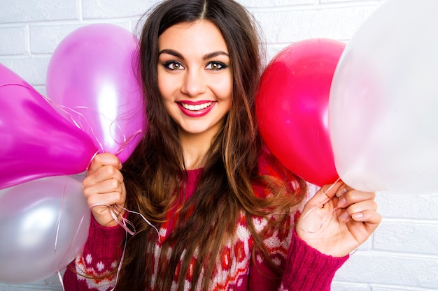 Helles lustiges Lebensstilbild zur hübschen jungen Frau im lässigen trendigen Pullover, der Spaß hat und Partyballons hält.