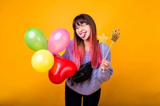 Helles lustiges Porträt der fröhlichen Hipster-Frau mit hellrosa Haaren, kuscheligem Pullover tragend, gefälschte Partykrone haltend und lächelnd, bereit für Party, gelbe Wand.