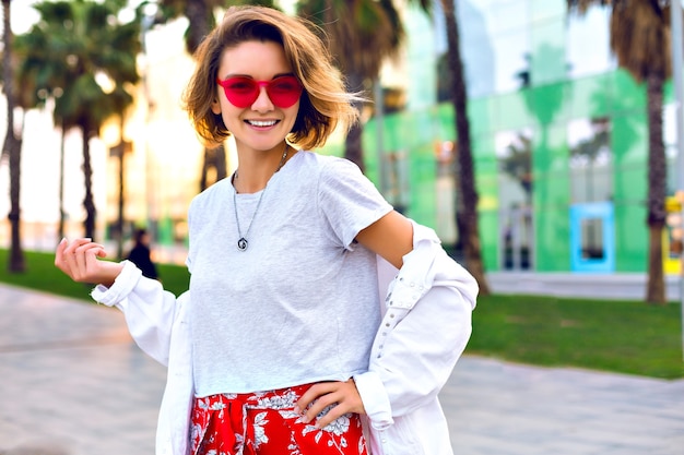Helles Porträt der Sommeraußenmode der stilvollen trendigen lächelnden Frau, die das stilvolle Hipster-Outfit, die weiße Jeansjacke und die Neon-Sonnenbrille trägt, Palmen herum, glückliche Reisestimmung.