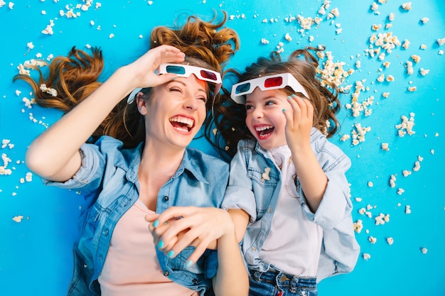 Helles stilvolles Bild von oben aufgeregter Mutter und Tochter, die auf blauem Boden in Popcorn liegen und in 3D-Gläsern lachen. Glückliche Familienzeit, Unterhaltung hübsche Mutter mit Kind, Glück ausdrücken