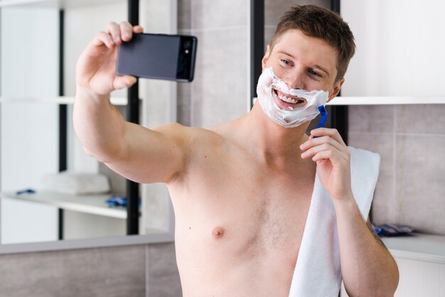 Hemdloser junger Mann, der im Badezimmer nimmt selfie auf Smartphone sich rasiert