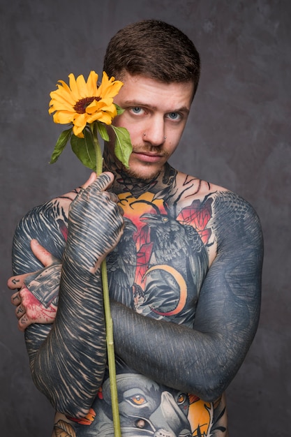 Hemdloser junger Mann mit Tätowierung auf seiner Karosserie, die in der Hand Sonnenblume gegen grauen Hintergrund hält