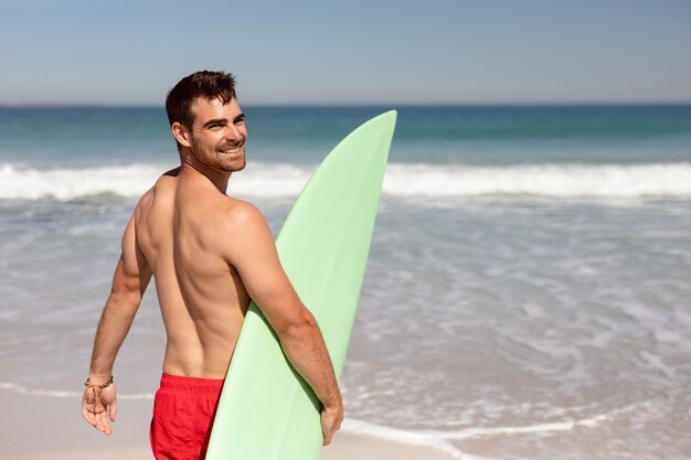 Hemdloser Mann mit dem Surfbrett, das Kamera auf Strand im Sonnenschein betrachtet