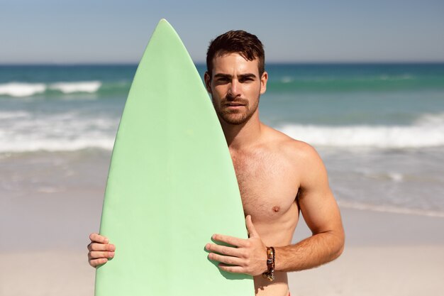 Hemdloser Mann mit dem Surfbrett, das Kamera auf Strand im Sonnenschein betrachtet