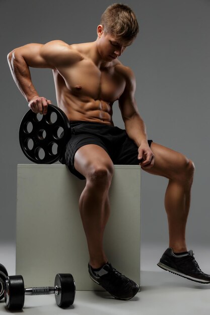 Hemdloser muskulöser Kerl sitzt auf weißer Kiste und hält Langhantelgewicht.