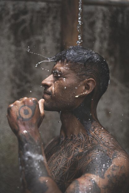 Hemdloser, muskulöser lateinamerikanischer Mann mit Tätowierungen, der nach dem Training auf dem Hintergrund der tropischen Blätter im Freien duscht. Dusche in der privaten Villa.