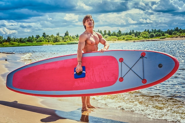 Hemdloser Surfer mit muskulösem Körper und seinem Surfbrett am Strand.