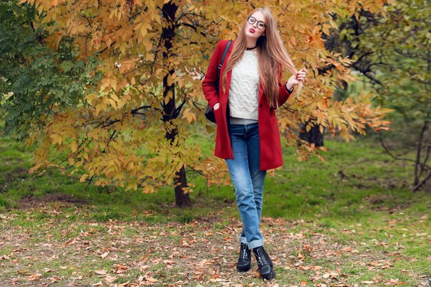 Herbstmodebild der vollen Höhe der hübschen netten Frau im roten stilvollen Mantel und in der Strickmütze, rote Lippen, die auf gelbem Park aufwerfen.