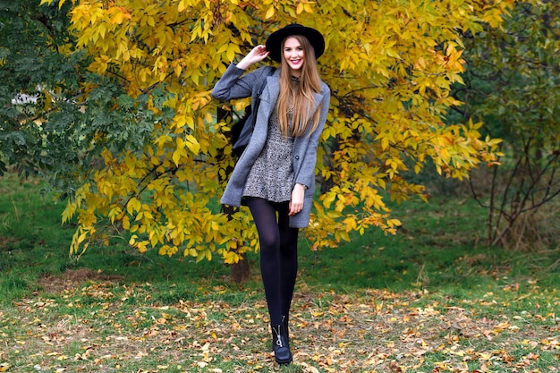 Herbstmodeporträt der eleganten Glamourfrau, die im erstaunlichen Stadtpark, im stilvollen Mantel, im Rucksack und im Weinlesehut aufwirft. Allein gehen, kaltes Wetter