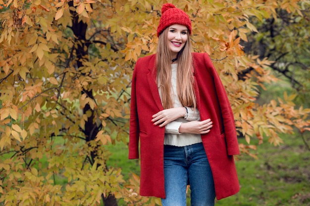 Herbstmodeporträt der glücklichen blonden Frau im roten stilvollen Mantel und in der Strickmütze, die im Park gehen.