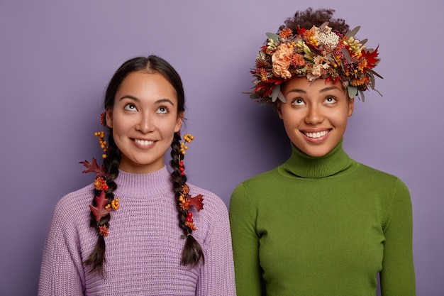 Herbstzeitkonzept. Fröhliche junge gemischtrassige Frauen in Freizeitkleidung, die sich oben konzentrieren, haben ein zahniges Lächeln, tragen Herbstblätter und Beeren im Haar, freuen sich über Herbstrabatte, posieren im Haus