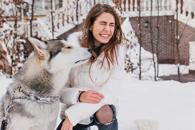 Herrliche Frau im weißen Kittel, die Winterspaziergang mit ihrem lustigen Hund genießt. Außenporträt der reizenden europäischen Frau, die mit Husky am schneebedeckten Hof spielt.