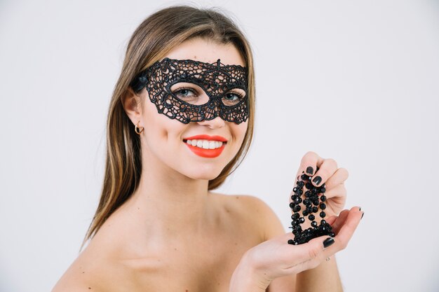 Herrliche lächelnde Frau in der schwarzen Karnevalsmaske, die schwarze Perlenhalskette hält