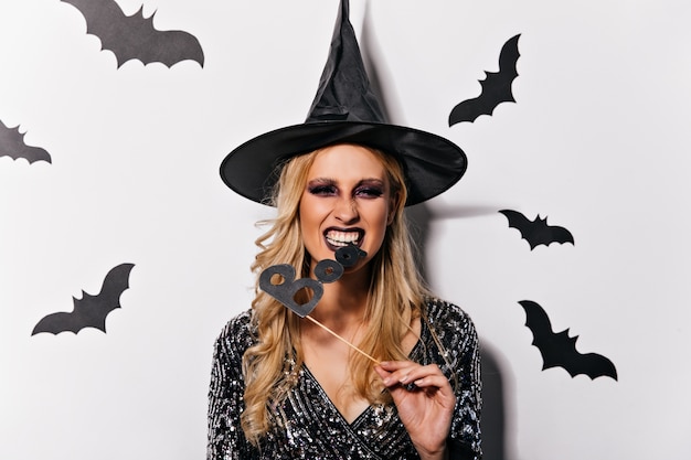 Hexe mit gewellter Frisur, die Halloween feiert. Ekstatischer weiblicher Vampir im lustigen Hut, der mit Fledermäusen aufwirft.