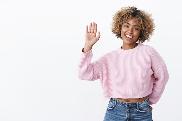 Hey, gib mir High Five. Freundliche, fröhliche, charismatische Freundin mit blondem Afro-Haarschnitt im Winter stylischer Pullover, der Palmen anhebt, grüßen Freund oder winken fröhlich und süß in die Kamera über weißer Wand