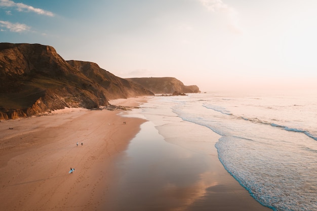 High Angle Shot der Meereswellen, die neben felsigen Klippen unter dem hellen Himmel zum Strand kommen