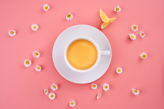 High Angle Shot einer Tasse Orangensaft, umgeben von kleinen Gänseblümchenblumen auf einer rosa Oberfläche