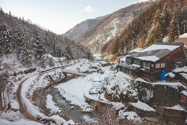 High Angle Shot eines Holzhauses, umgeben von bewaldeten Bergen, die im Winter mit Schnee bedeckt sind