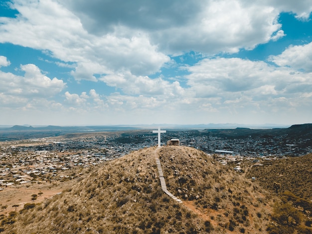 High Angle Shot von Hügeln bedeckt in trockenem Gras mit einem großen weißen Kreuz auf der Spitze unter einem blauen Himmel