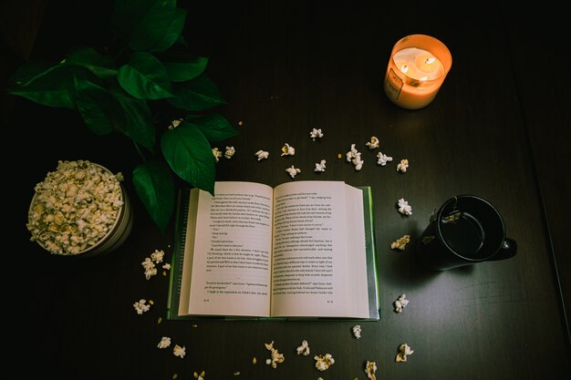 High Angle View eines offenen Buches und Popcorns auf dem Tisch mit einer brennenden Kerze und einer Tasse Tee
