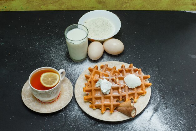 High Angle View Waffeln in Teller mit Tee, Eiern, Mehl, Milch auf Dunkelheit. horizontal