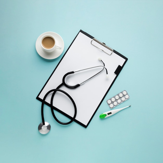 High Angle View Zwischenablage mit Papier und Kaffeetasse; Thermometer; Stethoskop; Blister verpackte Medizin gegen blauen Hintergrund