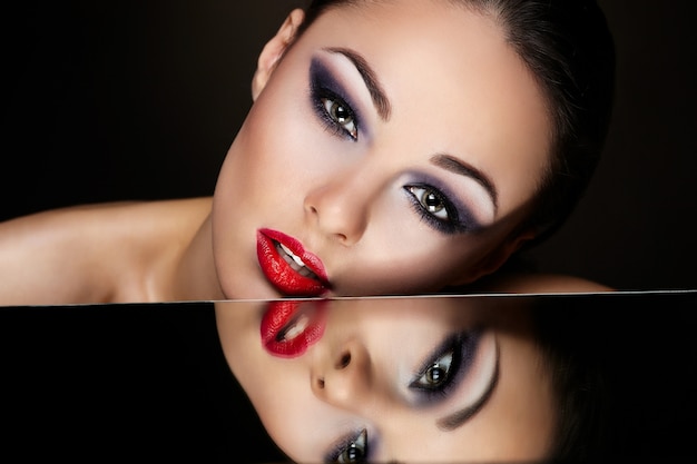 High Fashion Look. Glamour-Mode-Porträt des schönen sexy brünetten Mädchens mit hellem Make-up und roten Lippen und ihrem Spiegelbild im Spiegeltisch auf dunkel