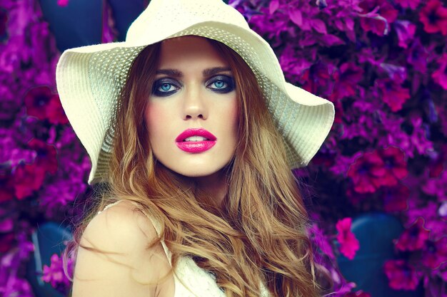 High Fashion Look.glamour Nahaufnahme Porträt des schönen sexy stilvollen blonden jungen Frau Modell mit hellem Make-up und rosa Lippen mit perfekter sauberer Haut im Hut nahe Sommerblumen